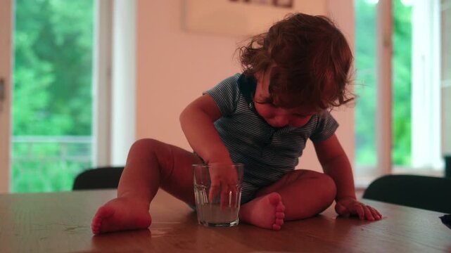 Toddler sitting on dining table dipping hand into drinking glass with curious expression, playful childhood moment expressing exploration mischief and everyday family life