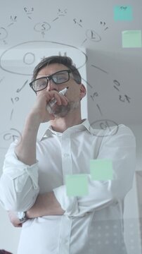 Thoughtful businessman wearing glasses concentrating on complex project plan. Analyzing strategy. Working with sticky notes and flowcharts on a clear glass board