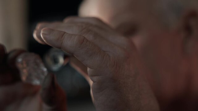 Elderly jeweler inspecting rough diamond with loupe at workbench, close up push in on hands face and gemstone.