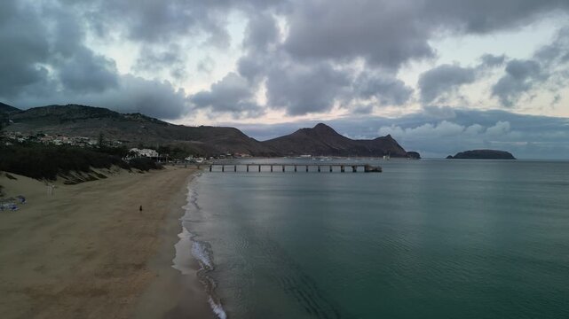Aerial drone view of long golden sand beach of Porto Santo Island, Madeira, Portugal. Cloudy sunset. Port and small marina. Mountains in the background