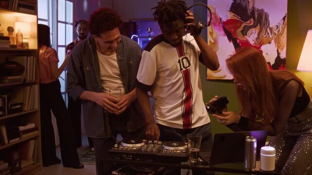 Medium full shot of young adult Black man playing music on DJ controller while cheerful friends observing and recording on vintage digital camera during home party