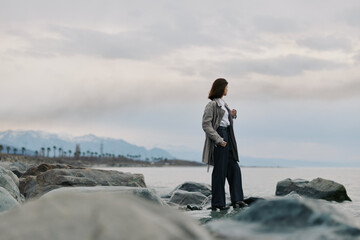 Woman in casual clothes stands on rocky shore looking at cloudy sky over calm sea. Concept of solitude, contemplation, and nature connection during overcast weather outdoors. © SHOTPRIME STUDIO