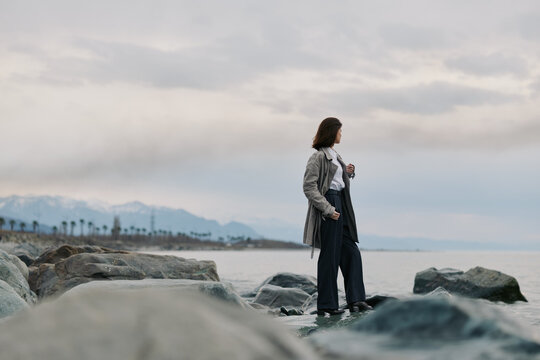 Woman in casual clothes stands on rocky shore looking at cloudy sky over calm sea. Concept of solitude, contemplation, and nature connection during overcast weather outdoors.