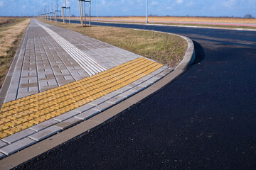 Fototapeta premium Yellow tactile paving for blind and visually impaired people on a new city sidewalk. Modern accessible urban environment with stone tiles and asphalt road. Concept of safety and inclusivity.