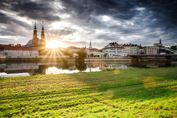 Malownicza panorama Opola o zachodzie słońca z widokiem na katedrę, rzekę Odrę i Most Piastowski. Słoneczne refleksy na wodzie i soczysta zieleń nadbrzeża tworzą spokojny, turystyczny klimat miasta. © Henryk Niestrój