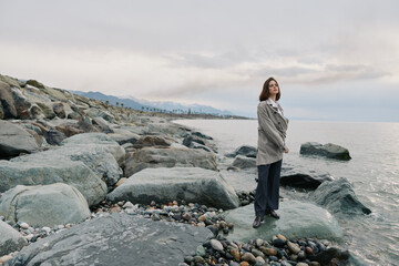Young woman in casual clothing stands on rocky beach near calm sea under cloudy sky. Natural...
