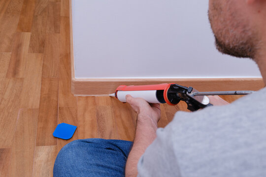 Man sealing the baseboard gap with a caulking gun on a natural iroko wood floor.
