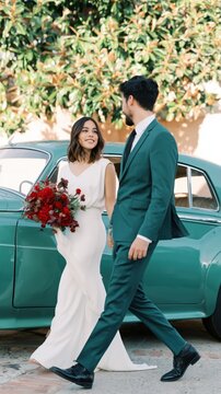 Caucasian female young bride in white dress and caucasian male young groom in teal suit walking by teal vintage car leafy background stylish wedding