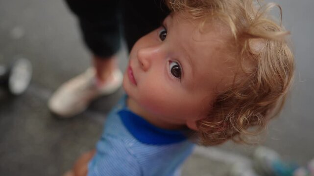 Toddler looking upward toward caregiver with wide curious eyes emotional childhood moment expressing trust attachment and search for reassurance in busy outdoor setting