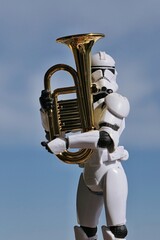 Naklejka premium Action figure of Star Wars clone trooper in white helmet and armor embracing model of some kind of euphonium brass instrument, blue spring skies with overcast clouds in background. 