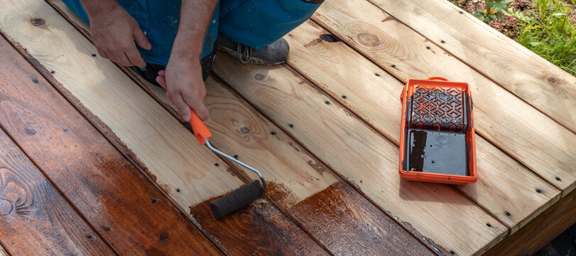 Worker applies wood stain with roller on outdoor deck