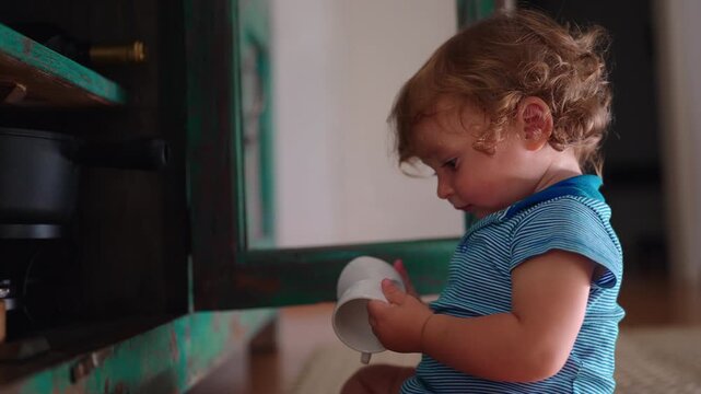 Toddler sitting on floor holding small cup while examining it with curiosity after taking objects from cabinet shelf, playful discovery moment inside family home