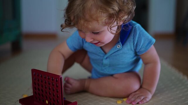Toddler sitting on play mat placing yellow token into red grid game while focusing carefully during quiet playtime at home, childhood curiosity learning and development