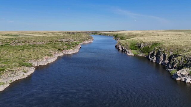 A scenic aerial shot from the middle of the Tobol River in the Kostanay region. The drone captures low rocky banks surrounded by vast green steppe on a bright summer day. The calm flow of water and th