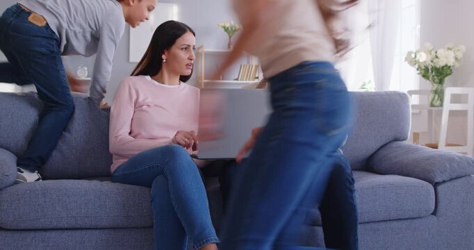 Smiling parents resting on sofa with laptop and discussing something while their active noisy children running and playing around them. Happy family enjoying leisure time and activities at home.