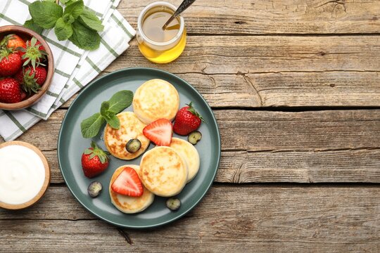 Delicious cottage cheese pancakes with berries, mint, sour cream and honey on wooden table, flat lay. Space for text