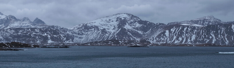 Panoramic view of snow-covered mountains across a Norwegian fjord under moody cloudy sky, capturing a cold Nordic winter seascape with calm ocean and dramatic landscape. © JEREMIE