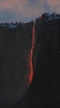 Vertical Screen: Yosemite National Park Firefall phenomenon where Horsetail Fall glows bright orange and red during sunset, cascading down El Capitan granite cliff with pink clouds above
