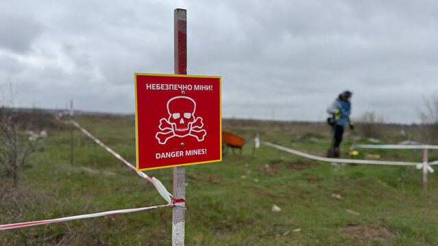 Red warning sign with skull symbol and Ukrainian text &ldquo;Danger Mines!&rdquo; marking a hazardous minefield area in Ukraine. In the background, a deminer conducts humanitarian mine clearance operations inside