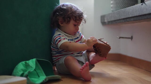 Toddler sitting on floor examining small shoe with focused attention while exploring everyday objects showing curiosity and early independence in home environment