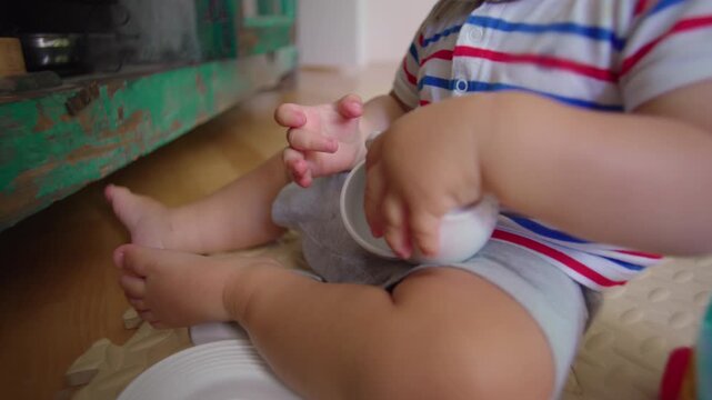Toddler sitting on floor holding small cups and kitchen items exploring objects with curiosity and tactile play during quiet everyday moment in home living space