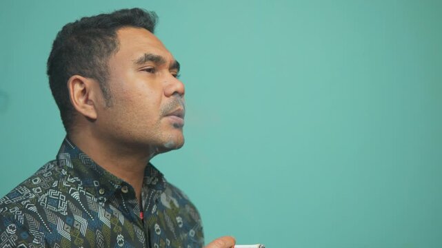 Side view of a man in a batik shirt smoking a cigarette. Concept of addiction, unhealthy lifestyle, and respiratory health issues.
