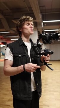 Vertical screen excited videographer smiles with expressive face holding camera on gimbal showing passion joy and satisfaction while enjoying creative filming process in gym