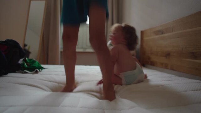 Toddler walking on mattress then falling softly before watching sibling jump nearby, capturing playful unpredictability, learning balance, and curious observation in authentic family moment