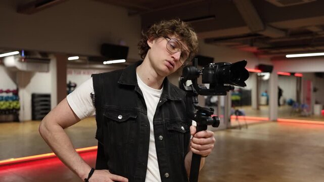 Cameraman with professional camera on gimbal looks exhausted and bored holding equipment and pausing during filming process showing lack of energy and routine work fatigue