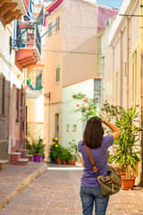 Fototapeta premium A woman photograph a narrow, cobblestone alley adorned with potted plants, including green leafy shrubs and blooming flowers. Carloforte Sardinia, Italy