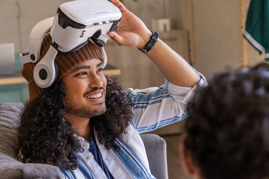 Diverse friends watching man with beanie lifting white VR headset while smiling on couch