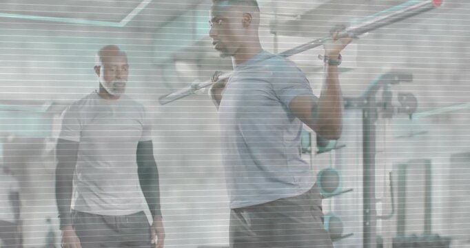 Vertical video: Gripping barbell, man doing squats at gym wearing blue tee, coach watching for form