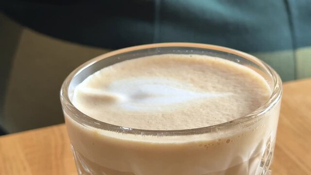 view of cappuccino in glass cup as teaspoon gently lifts white milk foam, showing creamy texture and warmth. Coffee ritual, cafe atmosphere, minimal composition, focus on foam detail.