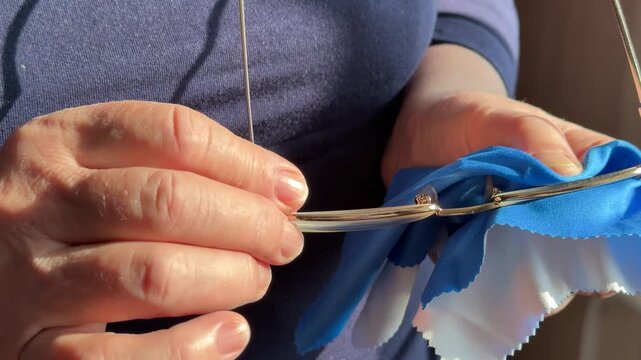 Closeup woman cleaning eyeglasses with blue microfiber cloth, sunlight catching gold frames and hands, slow deliberate wiping.