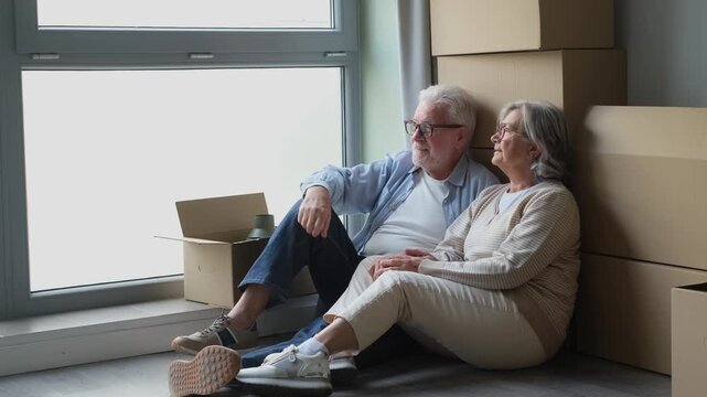 A happy senior couple sits on the floor of their new home, surrounded by moving boxes, discussing future plans and contemplating the next chapter of their lives together.