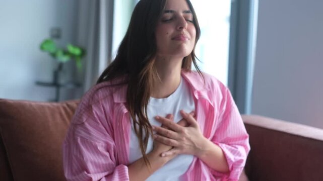 A young woman suffers from chest pain and shortness of breath while sitting on a sofa at home, indicating potential health issues or anxiety