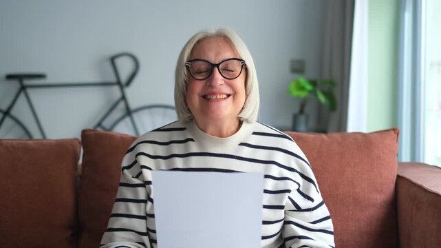 An older woman sits comfortably on a sofa at home, reading a document or letter. Her initial serious expression transforms into a joyful smile and laughter, suggesting positive news or happy memories.