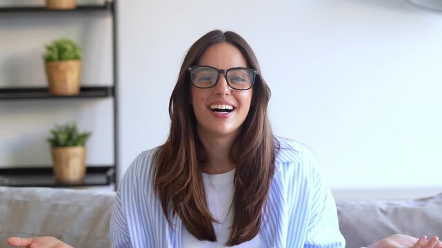 A cheerful young woman with glasses is actively talking and gesturing during a video call or online presentation from her modern home. Her expressive communication conveys happiness and confidence