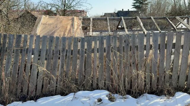 Weathered fence and broken greenhouse. Sunlit late winter scene with patchy snow, dried weeds tangled at base of leaning wooden.