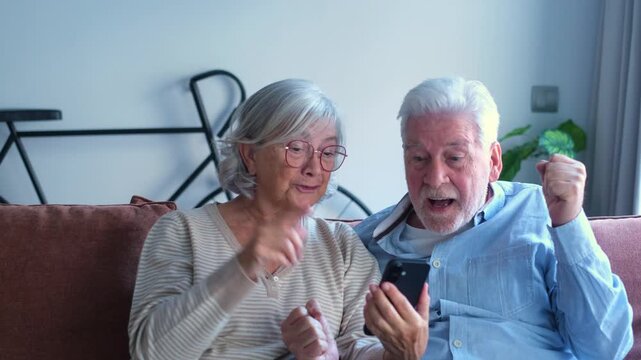 A joyful senior couple shares a moment of triumph while viewing content on a smartphone. They express excitement and happiness, celebrating good news together in their comfortable home.