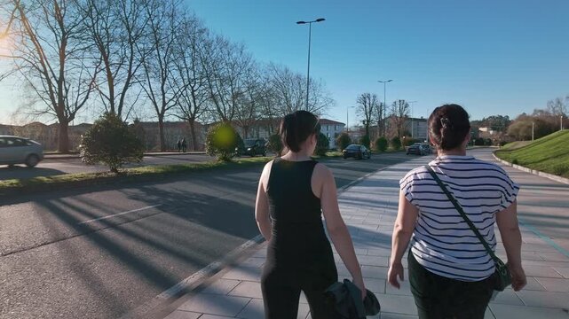 Following shot of a mother and daughter walking together along an urban section of the Camino de Santiago, engaging in conversation and enjoying a sunny day outdoors with leafless trees