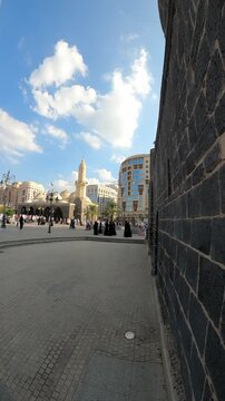 Wide-angle view panning from a historic dark stone wall to reveal pilgrims walking in the paved plaza near the beautiful Masjid Al Ghamamah in Madinah, Saudi Arabia under a bright blue sky.