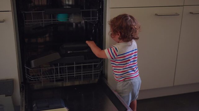 Toddler standing at open dishwasher reaching inside exploring kitchen appliance curious childhood moment of interaction with household objects during everyday routine at home