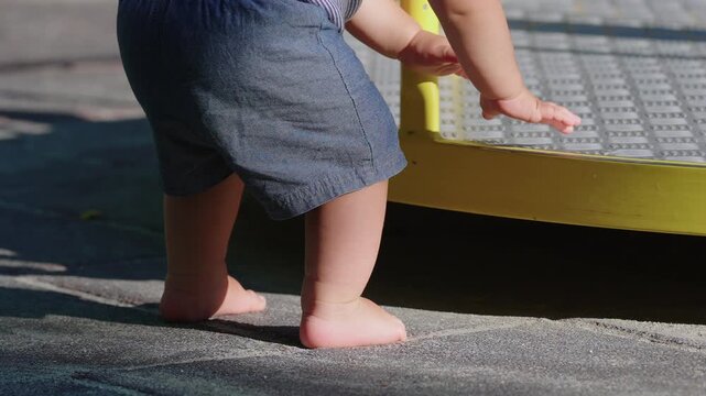 Barefoot toddler reaching toward playground merry go round then losing balance and falling to seated position capturing trial and error resilience during outdoor exploration