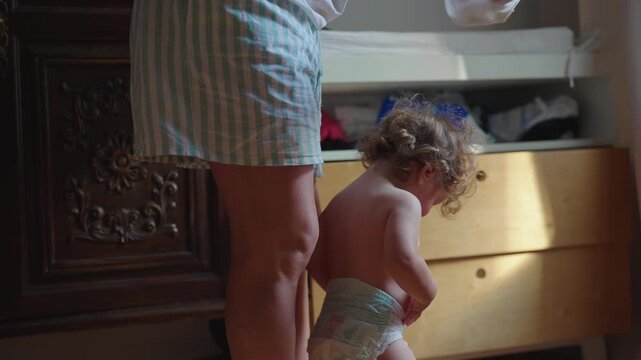 Mother helping toddler remove shirt while standing in bedroom near dresser capturing intimate caregiving routine and early childhood independence moment in home environment