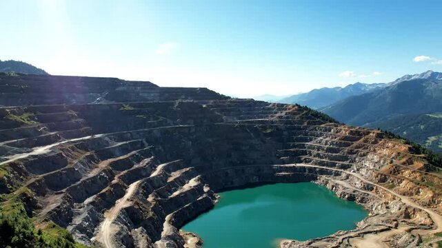 Aerial view of an open pit mine with a bright teal lake and mountain range under a clear blue sky