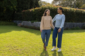 Obraz premium Diverse female friends walking together on grassy lawn near wall, wearing sweaters and jeans