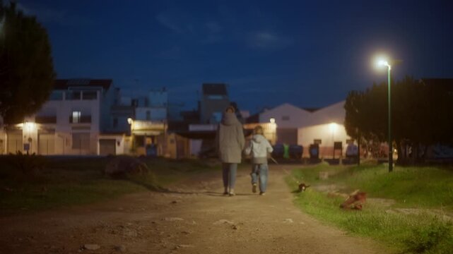 Family with woman and child walking a small dog on a dirt path at night near street lights