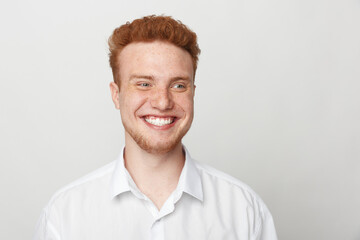 Obraz premium Smiling young man with red hair and beard wearing white shirt looks away on light background. Portrait of happy male model with natural expression and casual style in studio lighting.