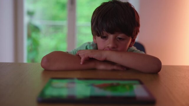 Boy resting head on arms looking at tablet on table thoughtful childhood moment of quiet screen time attention and passive engagement during everyday home routine